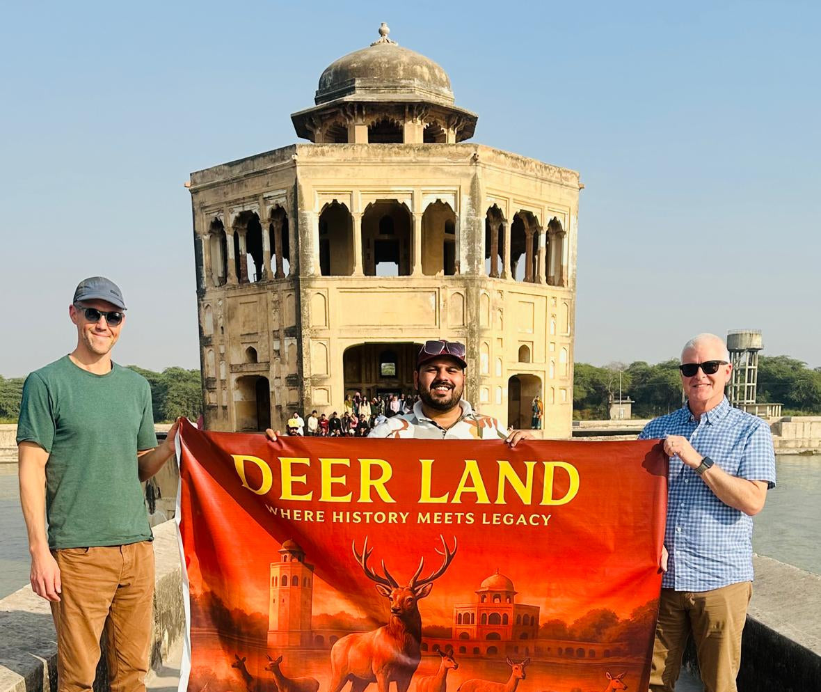 Three men holding a banner with 'DEER LAND' text and deer illustrations in front of a historical building.
